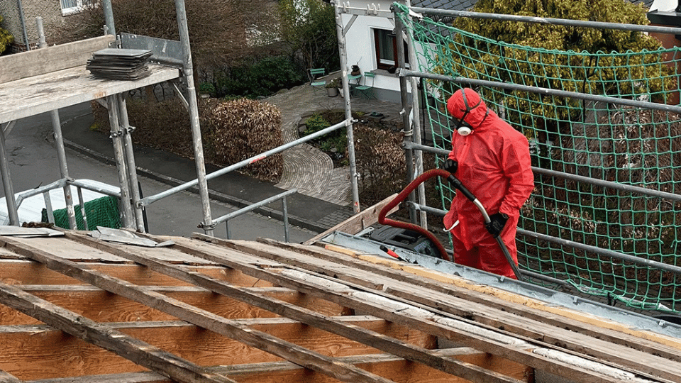 Asbestsanierung Mitarbeiter in rotem Schutzanzug bei der Asbestdach-Demontage in einem Wohngebiet